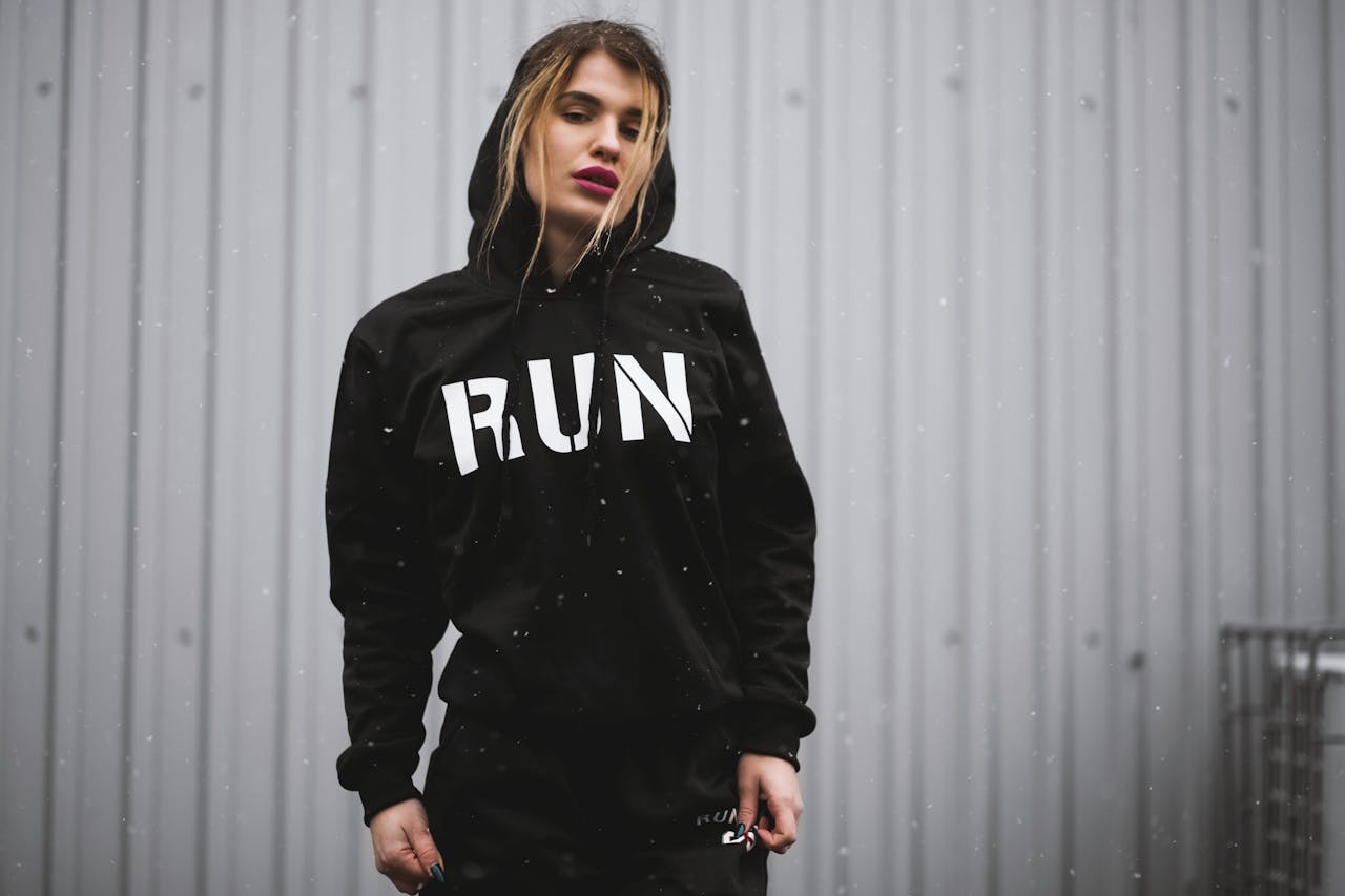 Home A woman in a black 'RUN' hoodie poses in an urban setting, exuding fashion and confidence.